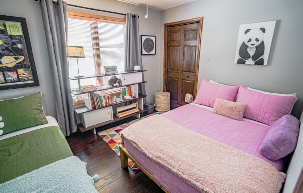 A colorful bedroom with a pink double bed, a green twin bed, and wooden shelving filled with books and decor.