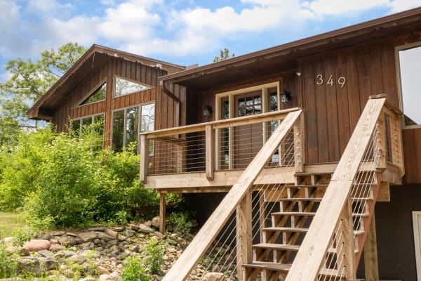 Exterior view of a wooden house with large windows and a front deck in a green landscape.