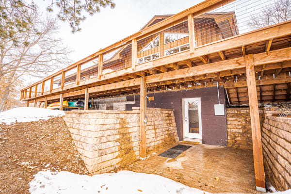Elevated wooden house with a deck above, stone walls below, and a snowy ground in winter.