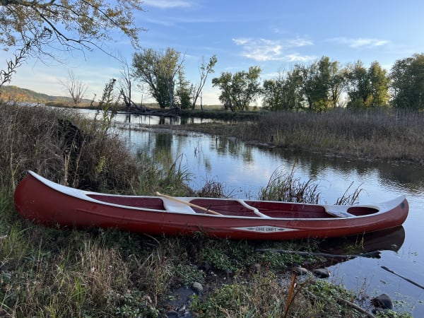 A red canoe on the grassy bank beside a calm river with trees in the background.