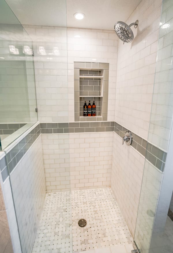 Interior view of a modern shower featuring white subway tiles, a built-in shelf with bottles, and a stone tiled floor.