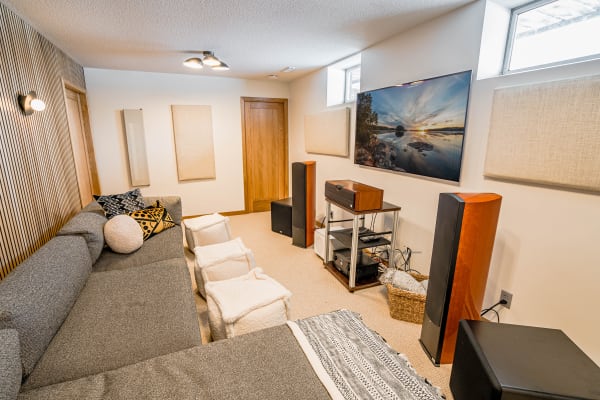 A cozy media room featuring a gray sectional sofa, decorative pillows, and a large TV displaying a scenic sunset. There are acoustic panels, a record player, and speakers present in the room.