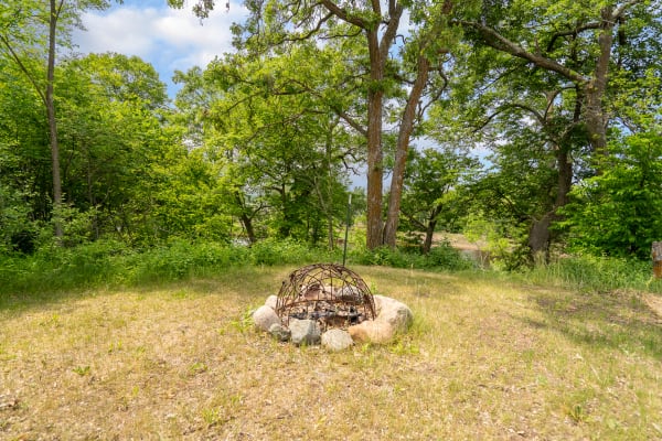 A fire pit made of stones and a metal grill set in a green landscape with trees.