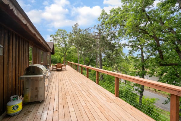 Spacious wooden deck adjacent to a cabin, featuring a grill and surrounded by trees.