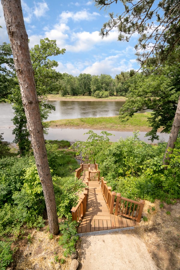 Wooden stairs descending through greenery to a calm riverbank under a blue sky.