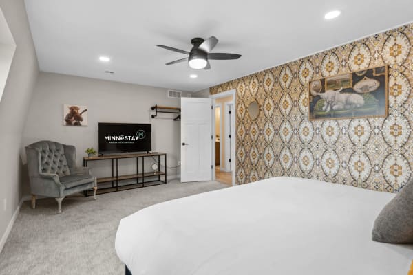 A cozy bedroom featuring patterned wallpaper, a bed with white bedding, a plush gray chair, and a sleek TV stand.