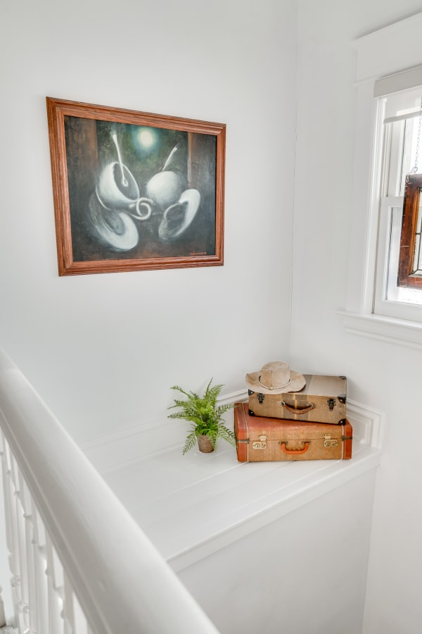 A corner with a framed abstract painting, vintage suitcases, a hat, and a small fern on a ledge.