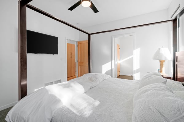 A spacious bedroom featuring a white quilted bed, mounted TV, and wooden furniture.