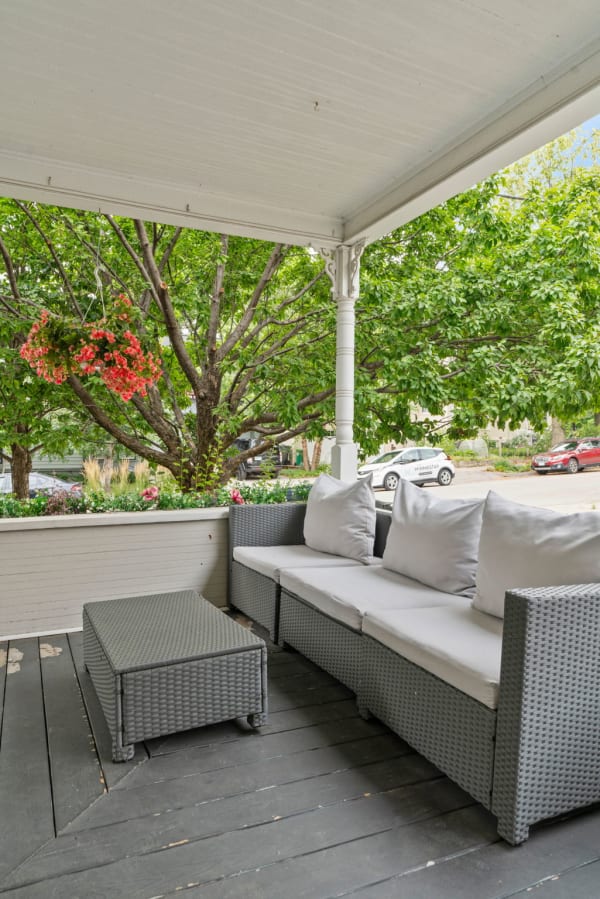 Cozy porch with gray wicker sofa, colorful flowers, and green trees.