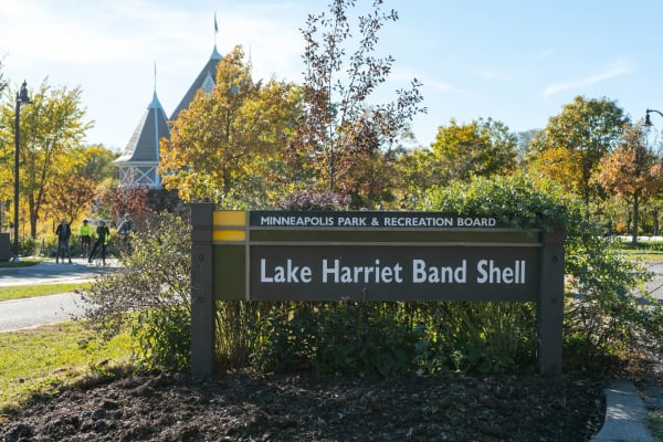 Sign for Lake Harriet Band Shell in a park with bicycles and pedestrians in the background.