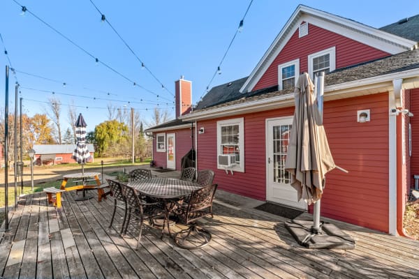 Outdoor deck with wooden flooring, a round table, chairs, and a red house in the background.