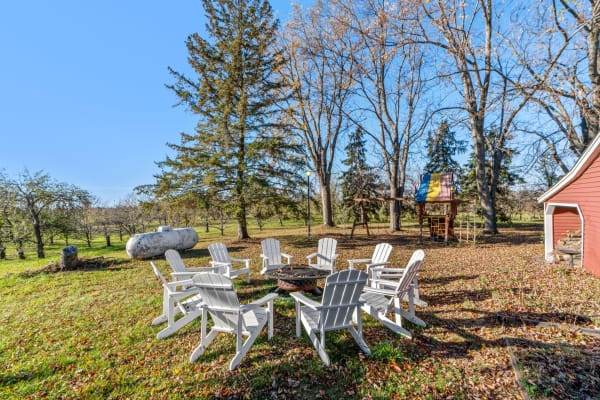 A circle of white Adirondack chairs around a fire pit, with a red barn and playset in the background.