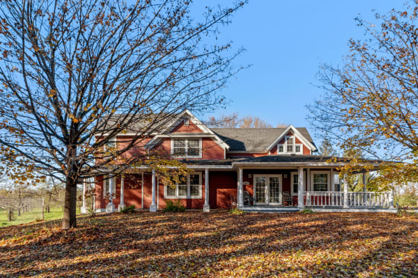A two-story red house with a porch in an autumn setting filled with fallen leaves.