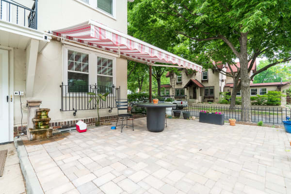 An outdoor patio featuring a round table, chairs, and a striped awning against a backdrop of greenery and traditional houses.