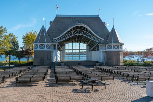An outdoor performance stage with benches arranged in front, surrounded by trees and water.
