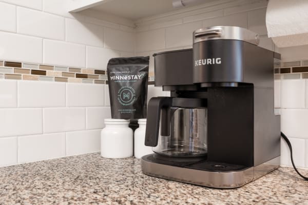 A black Keurig coffee maker on a granite countertop with a coffee bag and white jars.