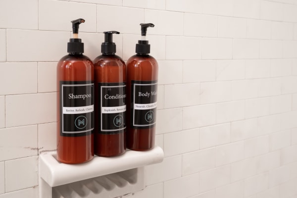 Three amber bottles labeled shampoo, conditioner, and body wash on a white shelf against white tiles.