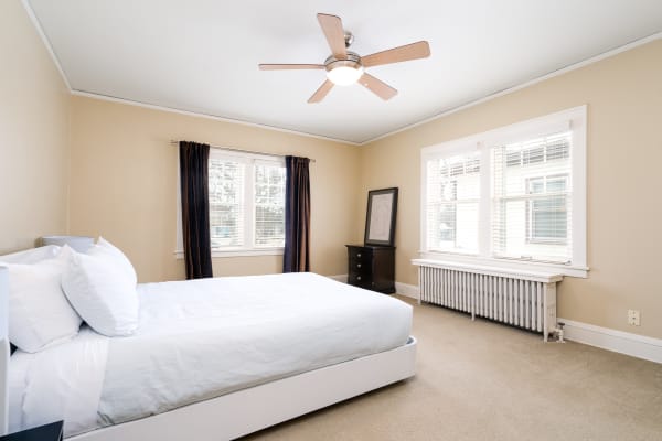A bright bedroom with a white platform bed, beige walls, dark curtains, and natural light from windows.