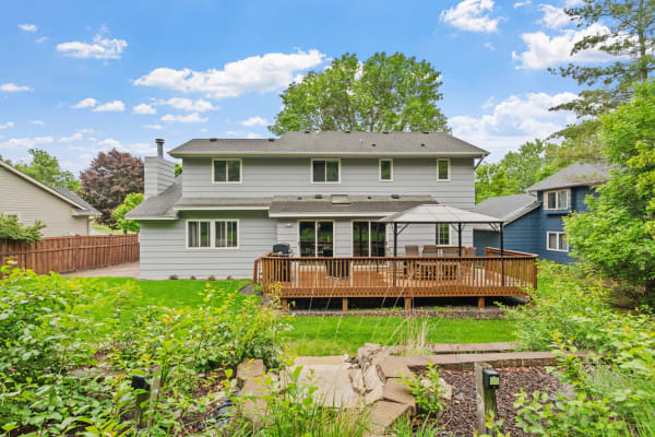 Backyard view of a two-story gray house with a wooden deck and green lawn under a blue sky.