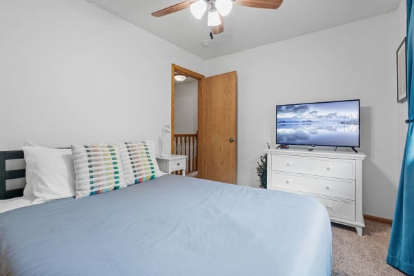 A cozy bedroom featuring a blue duvet, colorful striped pillows, a white dresser with a TV, and a wooden door.