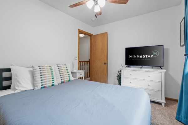 A modern bedroom with a light blue duvet and colorful pillows, featuring a MINNESTAY TV and white furniture.