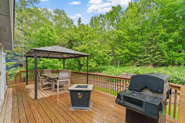 Wooden deck featuring a gazebo, dining set, and grill, surrounded by greenery.