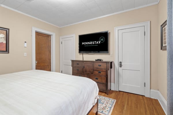 A warm-toned bedroom with a queen bed, wooden dresser, and wall-mounted TV.