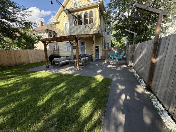 Backyard patio with a yellow house, wooden pergola, and teal chairs.