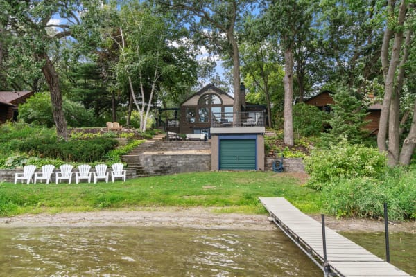 A cozy house by the lake with white Adirondack chairs, greenery, and a dock.