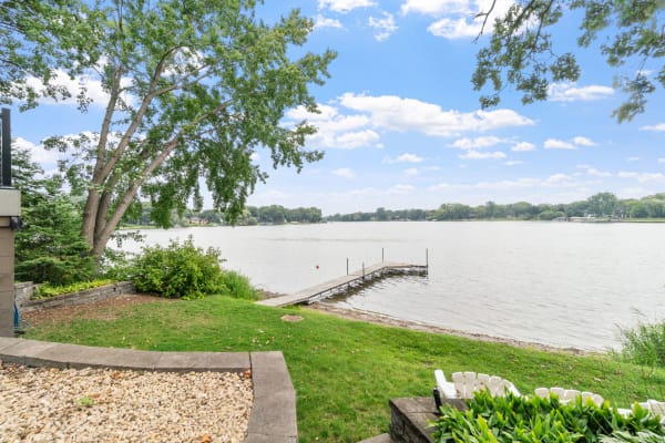 Lakeside view featuring a wooden dock, green lawn, and trees under a blue sky.
