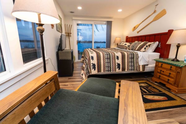 Lakeside bedroom featuring a red headboard bed, patterned quilt, green armchair, and sliding glass door.