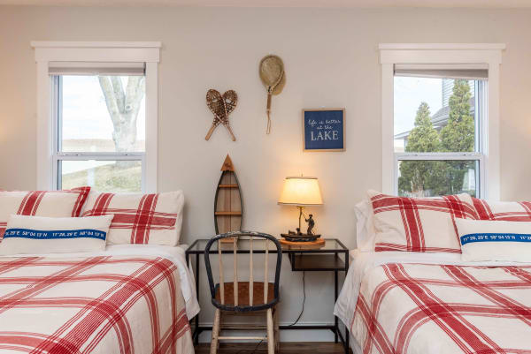 A cozy guest room with two twin beds featuring plaid bedding, a side table with a lamp, and lake-themed decor on the walls.