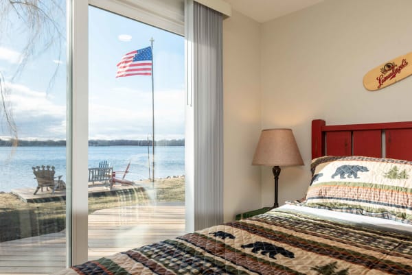 Cozy bedroom overlooking a lake with a flag and deck chairs outside.