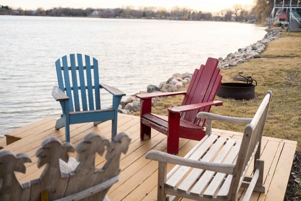 Three distinct chairs on a dock by a lake, surrounded by rocks and grass.