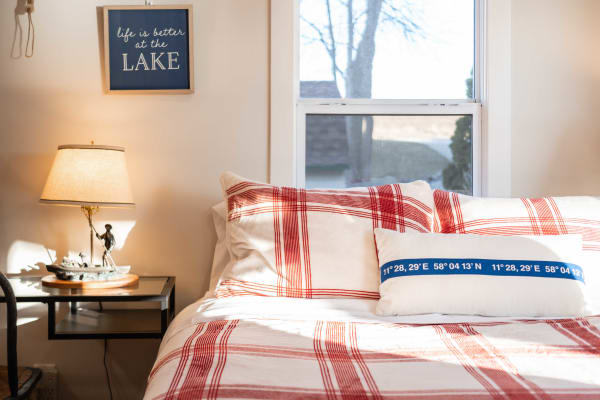 A bright bedroom featuring a red plaid bedspread, decorative pillows, and a lakeside sign.