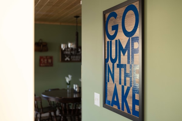 Wooden sign on a green wall stating 'GO JUMP IN THE LAKE' with a glimpse of a dining area in the background.