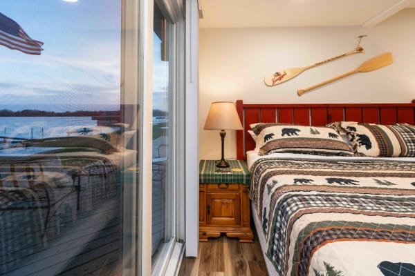A rustic bedroom featuring a red headboard, bear-patterned quilt, and a lakeside view through a sliding glass door.