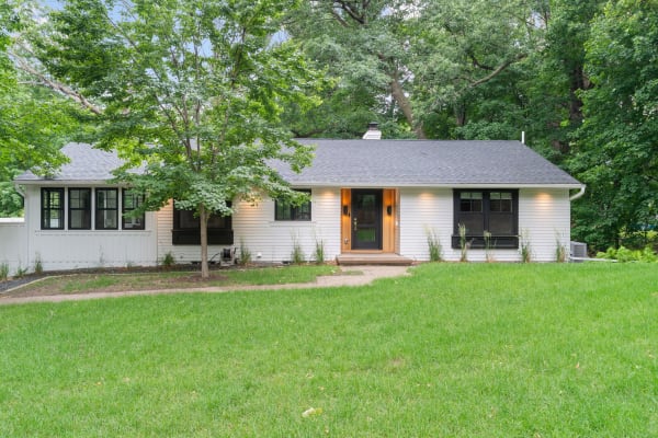 A modern white one-story home with a wooden door and black trimmed windows amidst green grass and trees.