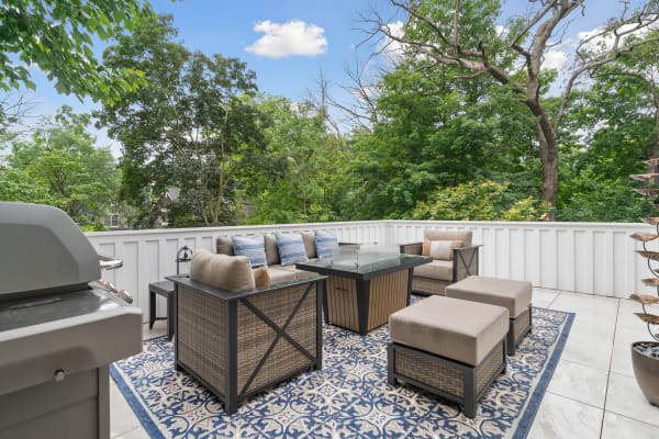 A modern outdoor seating area with a barbecue grill, greenery in the background, and a patterned rug.