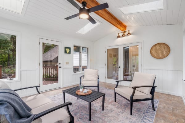 Sunroom with beige chairs, black table, and large windows showing green trees outside.