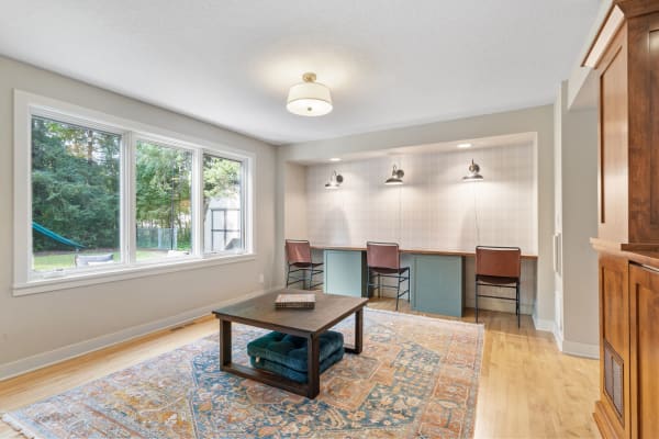 A cozy home office with large windows, a wooden table, and decorative rug.