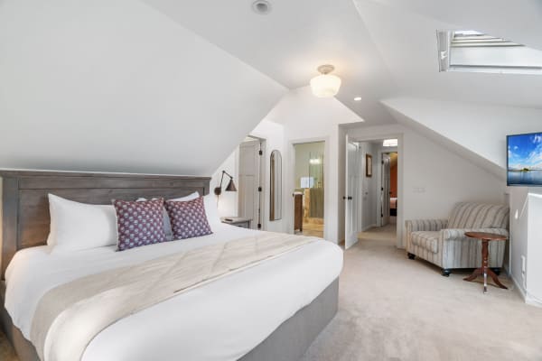 A modern, cozy bedroom with a queen bed, striped chair, and bathroom access in an attic space.