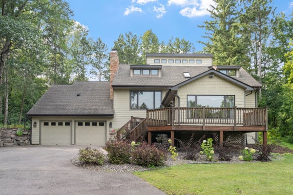 A charming two-story house with a wooden deck, surrounded by greenery and trees under a blue sky.