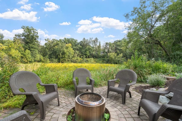 An outdoor seating area with wicker chairs around a fire pit, set against a lush green landscape.