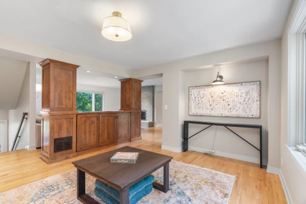 A spacious living room with wooden furniture, a dark coffee table on a patterned rug, and a floral artwork on the wall.