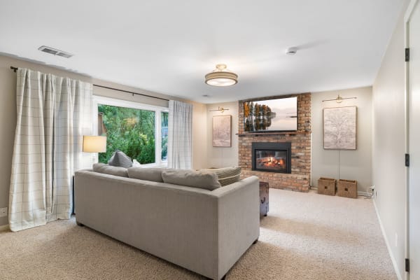 Living room featuring a grey sectional sofa, a brick fireplace with a mounted TV, curtains, and a view of greenery.