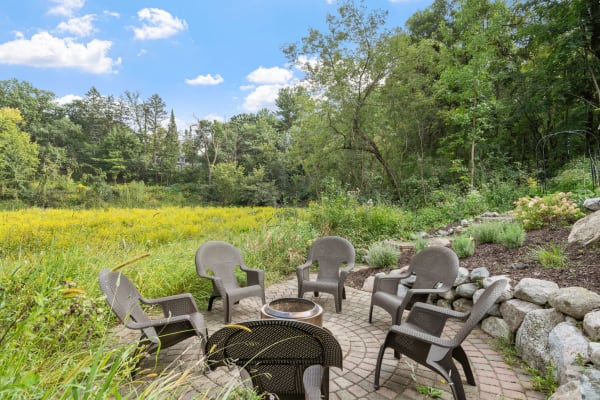 Outdoor seating area with chairs arranged around a fire pit, set against a backdrop of greenery and blue sky.