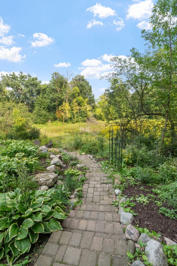 A cobblestone path in a garden leading to a green field, surrounded by plants and trees.