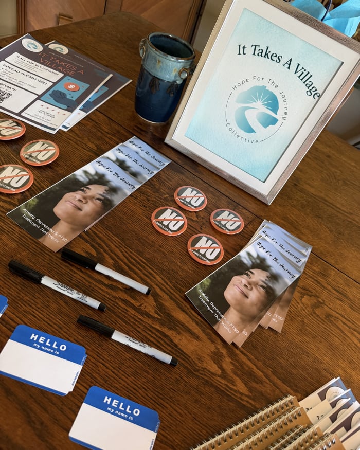 A Gatherings For Good welcome table with Hope For The Journey brochures, name tags, and an It Takes A Village sign