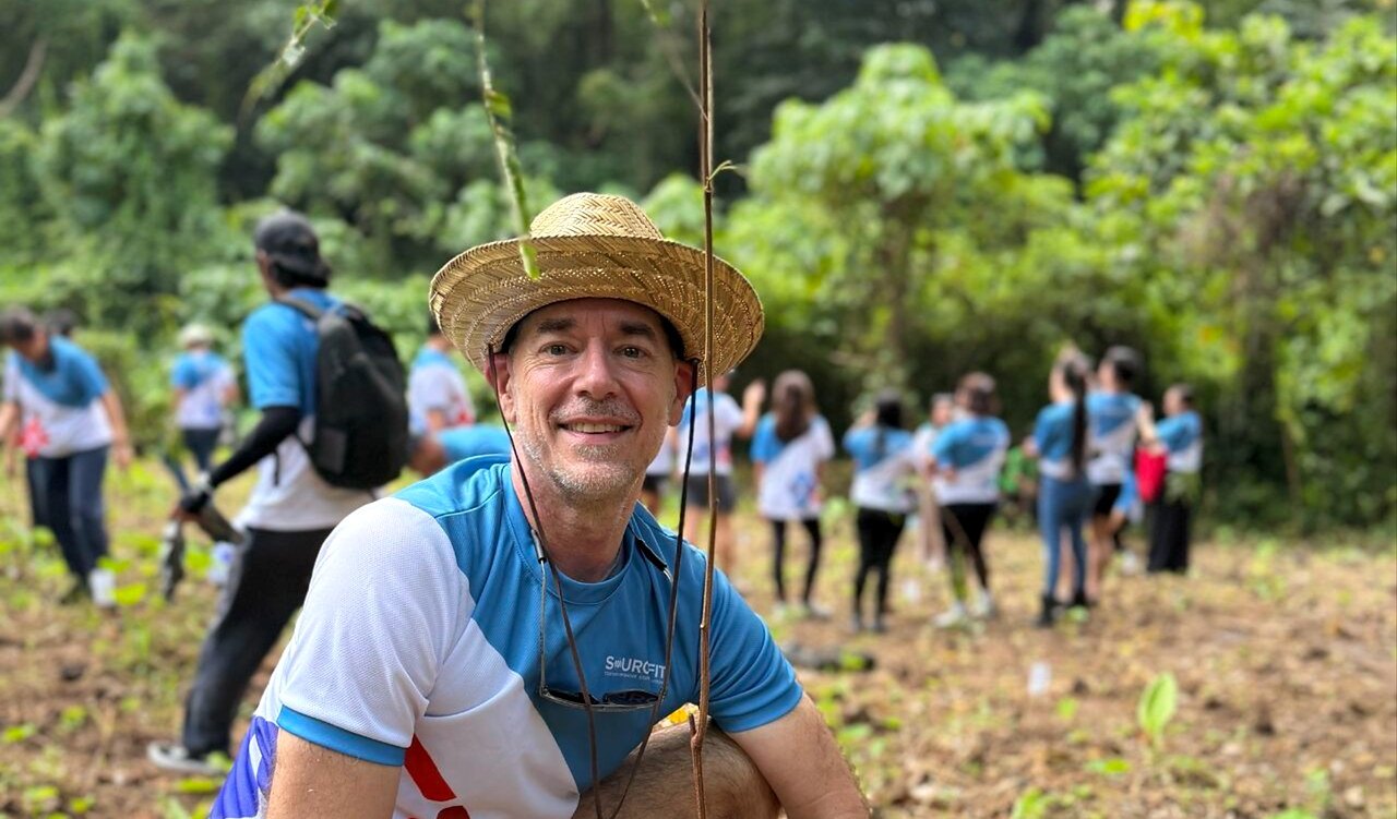 Andy Schachtel in a straw hat and Sourcefit shirt, smiling, with volunteers planting trees behind him